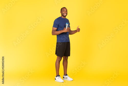 African Male Gesturing Thumbs Up Holding Fitness Bottle, Yellow Background © Prostock-studio