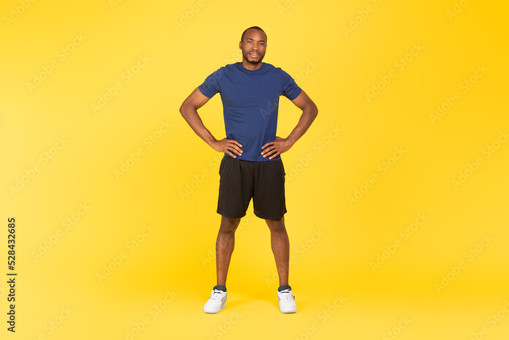 Sporty Black Male Holding Hands On Hips Standing, Yellow Background