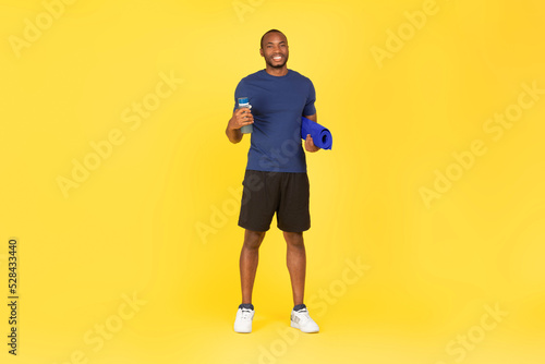 African Male Holding Water Bottle And Fitness Mat, Yellow Background © Prostock-studio