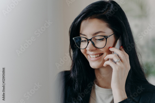 Happy woman lawyer keeps mobile phone near ear, has pleasant conversation with client, discuss working issues, smiles gently, wears transparent glasses, formal wear. Female employee calls someone