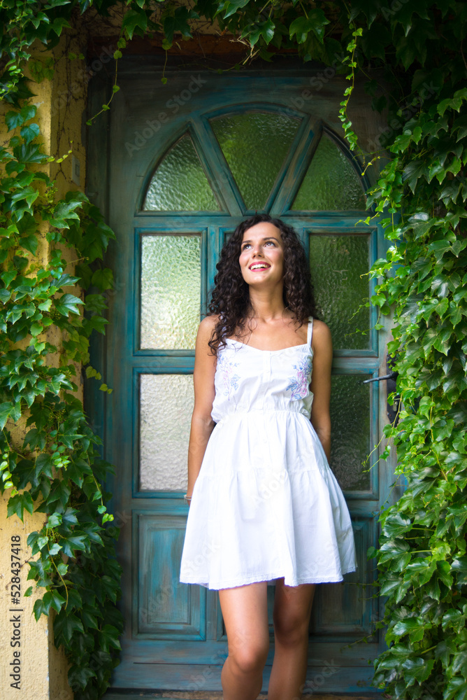 Young smiling elegant woman standing in front of home wooden door surrounded by ivy Young smiling elegant woman standing in front of home wooden door surrounded by ivy