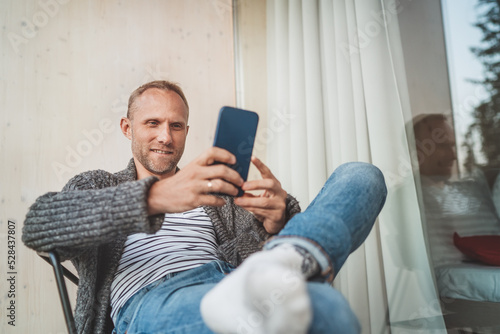 Smiling middle-aged man dressed open cardigan, jeans, and warm socks sitting on house balcony and smiling when he using modern smartphone. Everyday lifestyle photo with modern devices concept image..