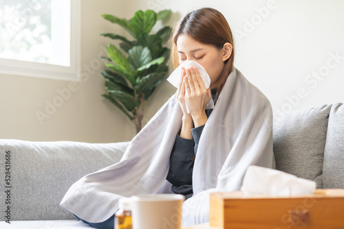 Quadro em tela Sick, influenza asian young woman have a fever, flu hand in use tissues paper sneezing runny nose covered body with blanket while sitting rest on sofa, couch take medicine at home