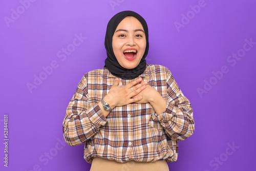 Amazed young Asian woman in plaid shirt hands on chest, looking at camera with wow face expression isolated on purple background