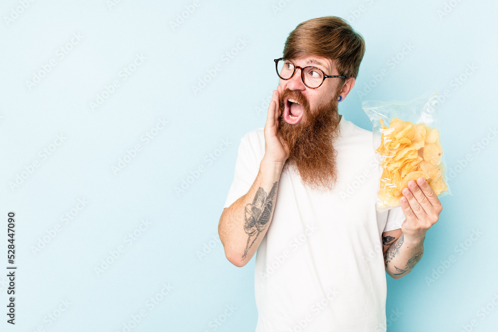 Young caucasian man holding a bag of chips isolated on blue background is saying a secret hot braking news and looking aside Young caucasian man holding a bag of chips isolated on blue background is saying a secret hot braking news and looking aside