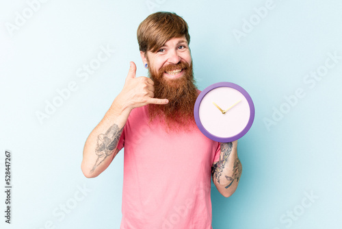 Young caucasian man holding a clock isolated on blue background showing a mobile phone call gesture with fingers. © Asier