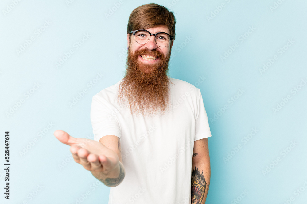 Young caucasian red-haired man isolated on blue background stretching hand at camera in greeting gesture. Young caucasian red-haired man isolated on blue background stretching hand at camera in greeting gesture.