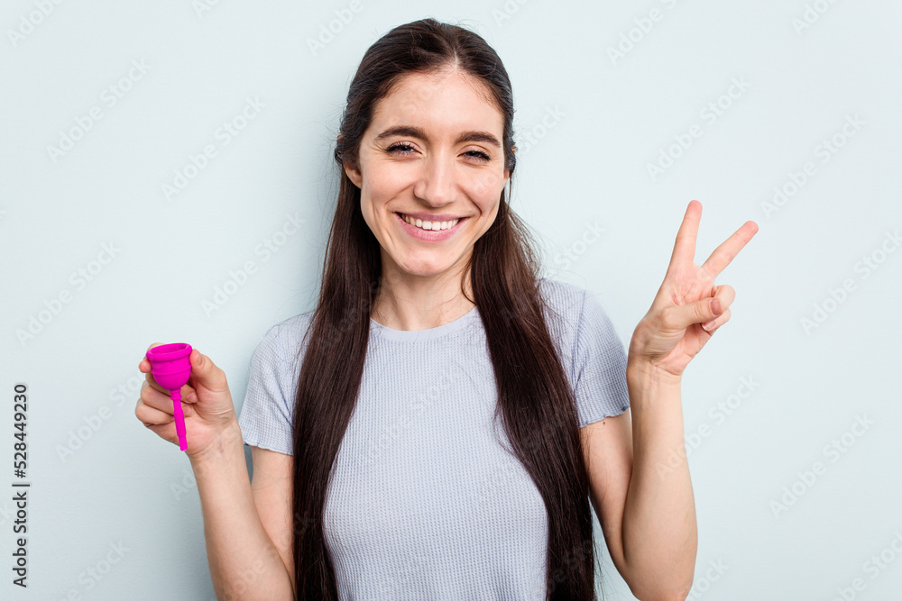 Young caucasian woman holding a sanitary napkin isolated on blue background joyful and carefree showing a peace symbol with fingers. Young caucasian woman holding a sanitary napkin isolated on blue background joyful and carefree showing a peace symbol with fingers.