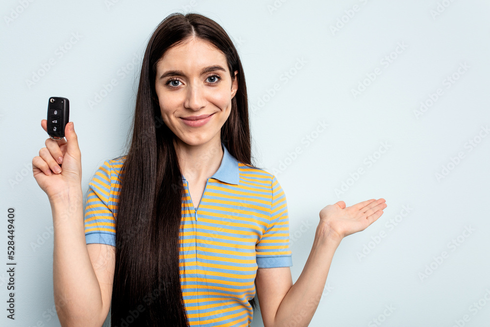 Young caucasian woman holding car keys isolated on blue background showing a copy space on a palm and holding another hand on waist. Young caucasian woman holding car keys isolated on blue background showing a copy space on a palm and holding another hand on waist.