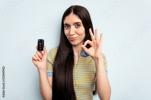 Young caucasian woman holding car keys isolated on blue background cheerful and confident showing ok gesture.