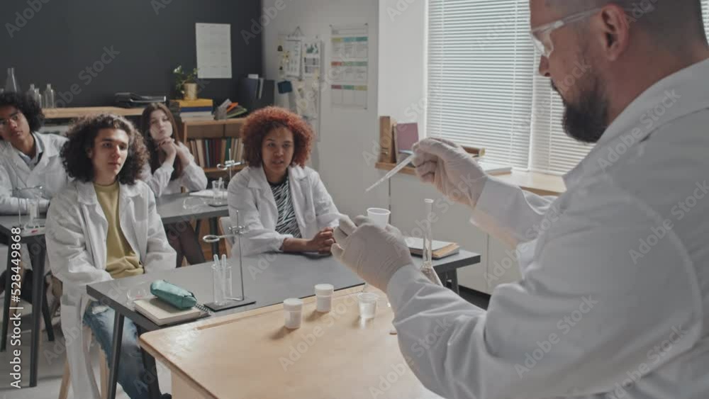 Over-shoulder of male chemistry teacher in medical gown and protective goggles standing in front of high school students in morning, showing experiment Over-shoulder of male chemistry teacher in medical gown and protective goggles standing in front of high school students in morning, showing experiment