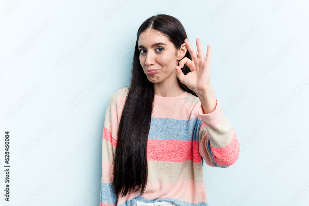 Young caucasian woman isolated on blue background winks an eye and holds an okay gesture with hand. Young caucasian woman isolated on blue background winks an eye and holds an okay gesture with hand.