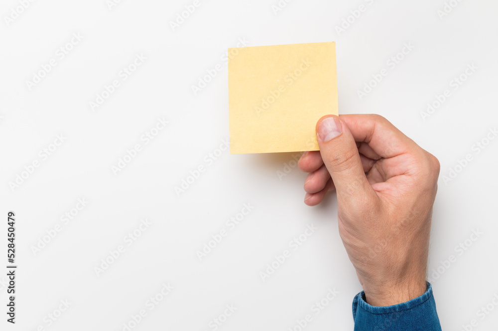 A man's hand in a denim shirt holds a stationery sticker paper for writing on a white background. Blank paper blank reminder to do list, office supplies note message, office and business concept. A man's hand in a denim shirt holds a stationery sticker paper for writing on a white background. Blank paper blank reminder to do list, office supplies note message, office and business concept.