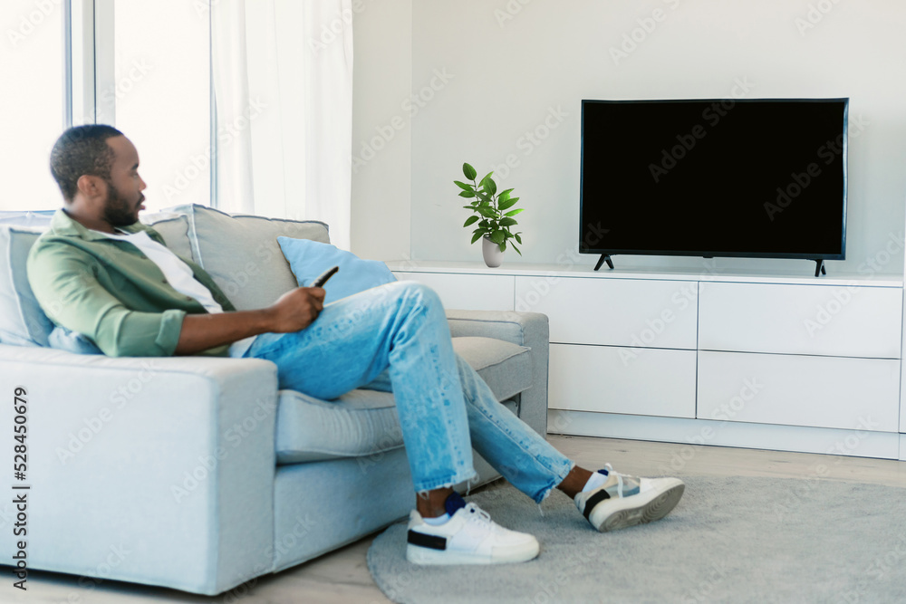 Young black man watching tv with empty screen, resting on sofa in living room interior, mockup