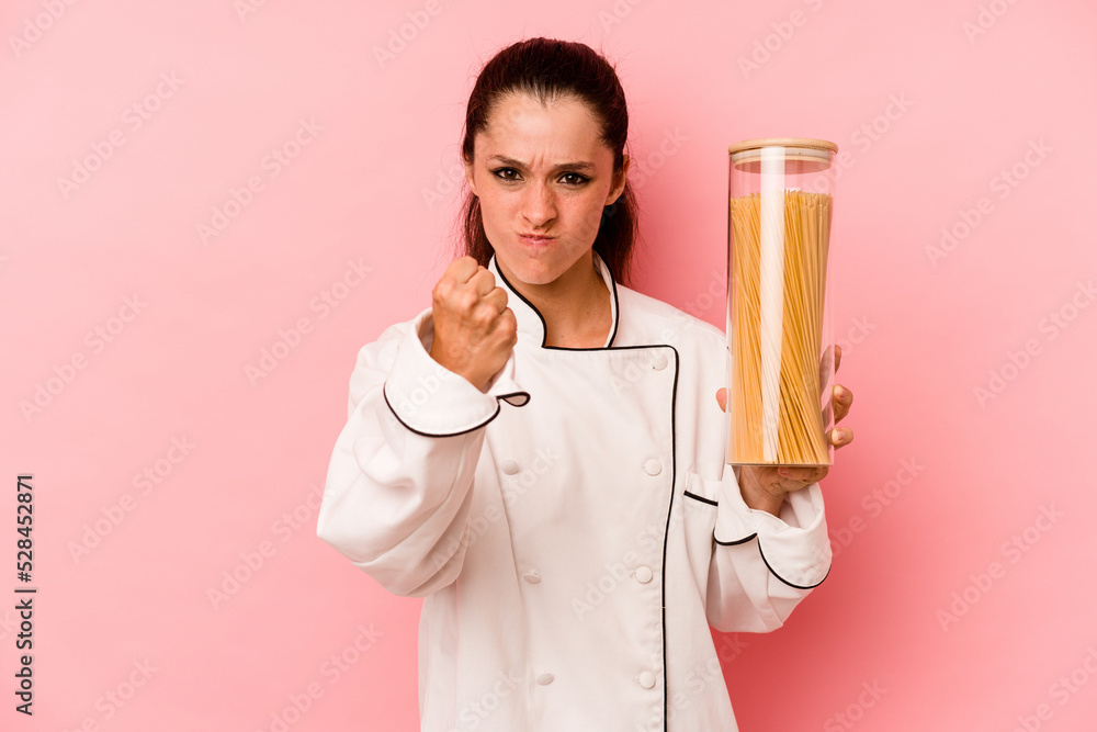 Young chef caucasian woman holding a spaghettis jar isolated on pink background showing fist to camera, aggressive facial expression. Young chef caucasian woman holding a spaghettis jar isolated on pink background showing fist to camera, aggressive facial expression.