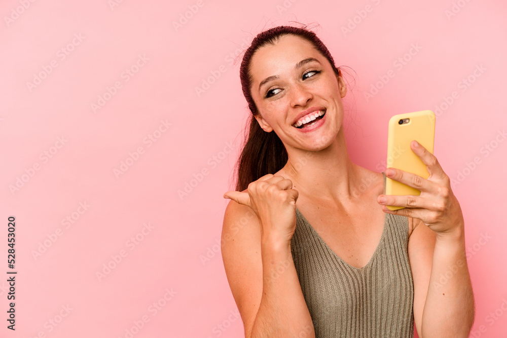 Young caucasian woman holding mobile phone isolated on pink background points with thumb finger away, laughing and carefree. Young caucasian woman holding mobile phone isolated on pink background points with thumb finger away, laughing and carefree.