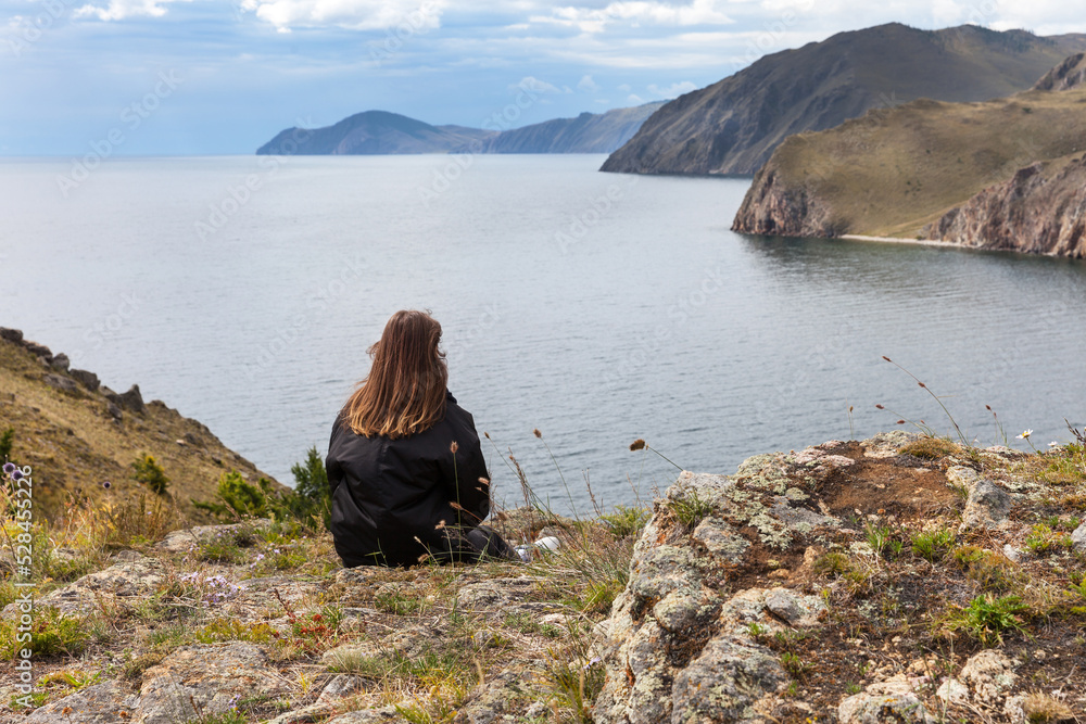 Fototapeta premium A tourist girl sits on a high rock and admires a beautiful view of the shore of Baikal Lake. Journey along the lake, outdoor recreation