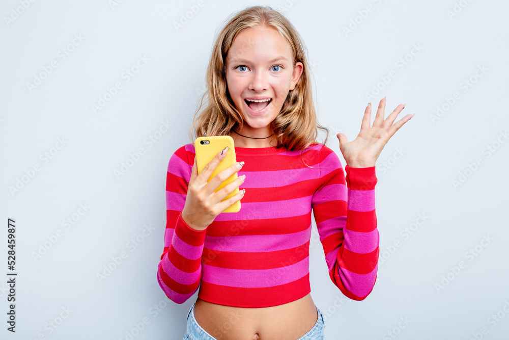 Caucasian teen girl holding mobile phone isolated on blue background receiving a pleasant surprise, excited and raising hands. Caucasian teen girl holding mobile phone isolated on blue background receiving a pleasant surprise, excited and raising hands.