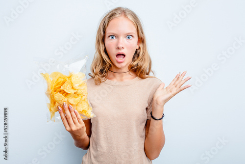 Caucasian teen girl holding bag of chips isolated on blue background surprised and shocked.
