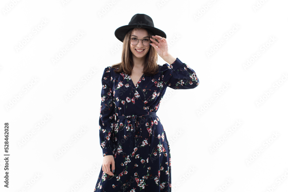 A brunette in a long floral dress with a hat poses for the camera in the studio A brunette in a long floral dress with a hat poses for the camera in the studio