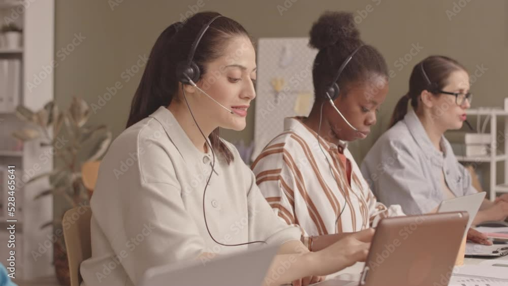 Waist up slowmo of young Middle Eastern woman with female colleagues working together at call center in office, talking to clients via headsets Waist up slowmo of young Middle Eastern woman with female colleagues working together at call center in office, talking to clients via headsets