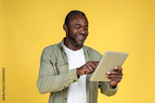 Cheerful handsome adult black man in casual typing on tablet, reading blog, isolated on yellow background photo