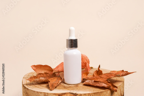 White glass cosmetic dropper bottle standing on wooden disk with dry leaves on beige background. Autumn composition. Concept of natural and organic skincare products presentation
