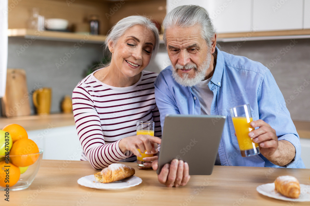 Cheerful Elderly Couple Having Breakfast Together And Using Digital Tablet In Kitchen Cheerful Elderly Couple Having Breakfast Together And Using Digital Tablet In Kitchen