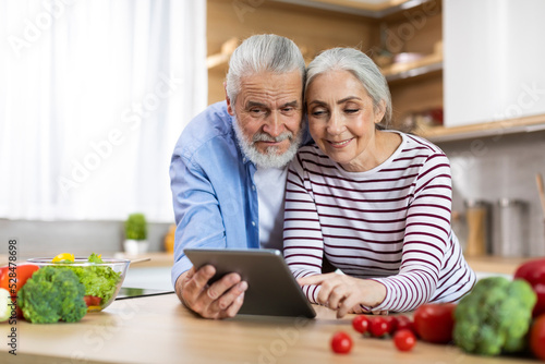 Portrait Of Happy Elderly Spouses Using Digital Tablet In Kitchen Interior,