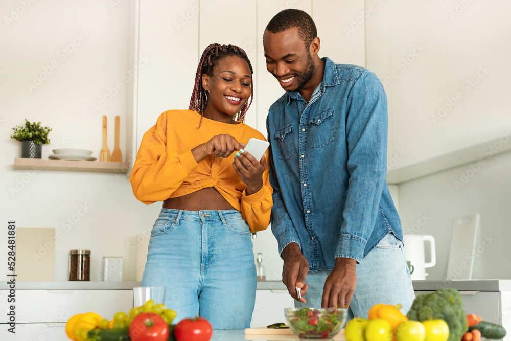 Loving black couple reading food blog on smartphone, cooking together healthy vegetable salad at home kitchen Loving black couple reading food blog on smartphone, cooking together healthy vegetable salad at home kitchen