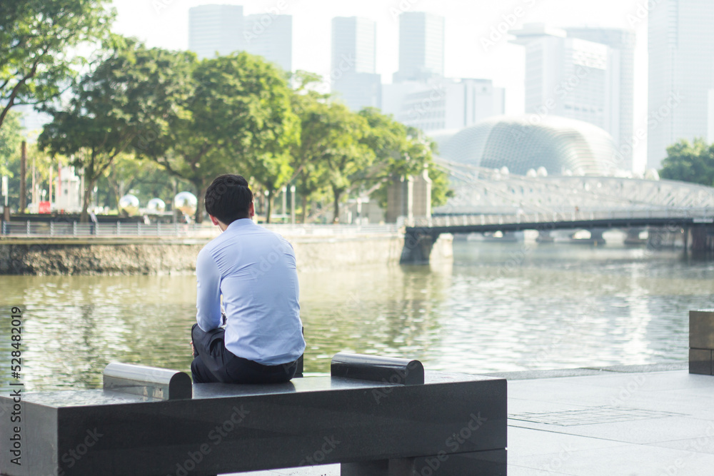 A man sits alone along side the river with the cityscape in the background. A man sits alone along side the river with the cityscape in the background.