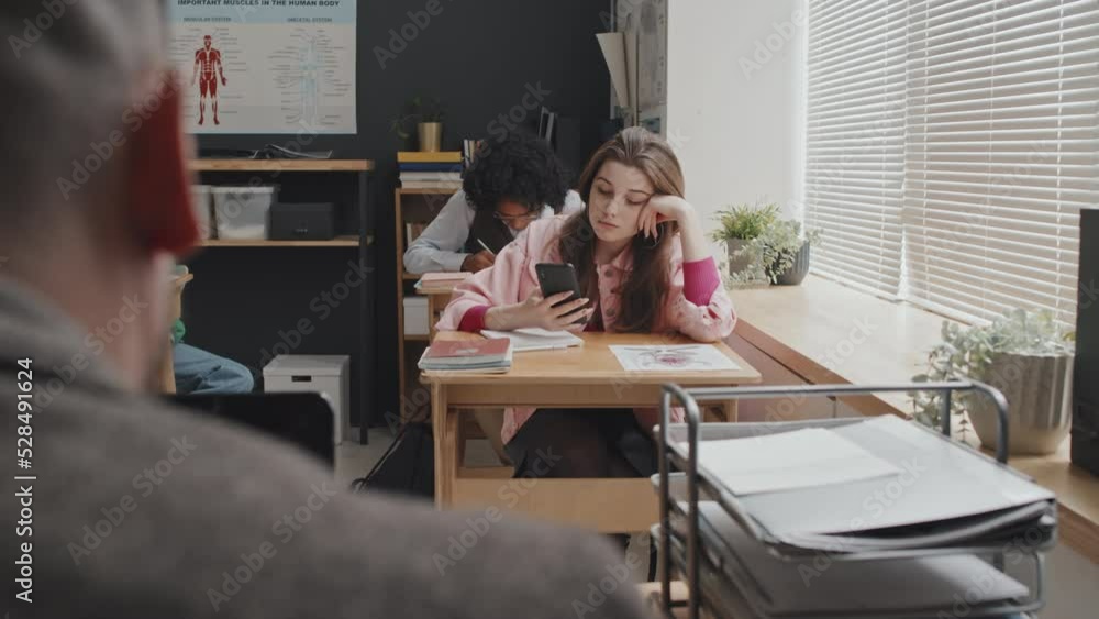 Medium long over-shoulder of unrecognizable teacher looking at female Caucasian high school student sitting at desk in class in morning, using smartphone Medium long over-shoulder of unrecognizable teacher looking at female Caucasian high school student sitting at desk in class in morning, using smartphone