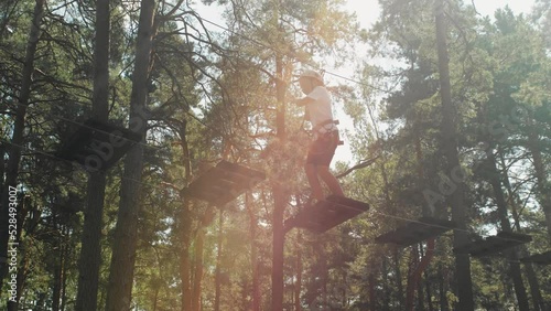 Brave teenage girl in gear and helmet, overcoming obstacles in a rope park in a sunny wooded area. Side view from below