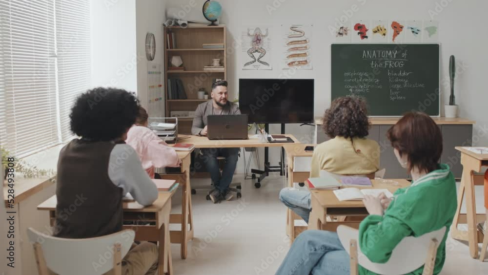 Rear tracking right of diverse high school students sitting at desks in classroom, talking to male teacher at daytime Rear tracking right of diverse high school students sitting at desks in classroom, talking to male teacher at daytime