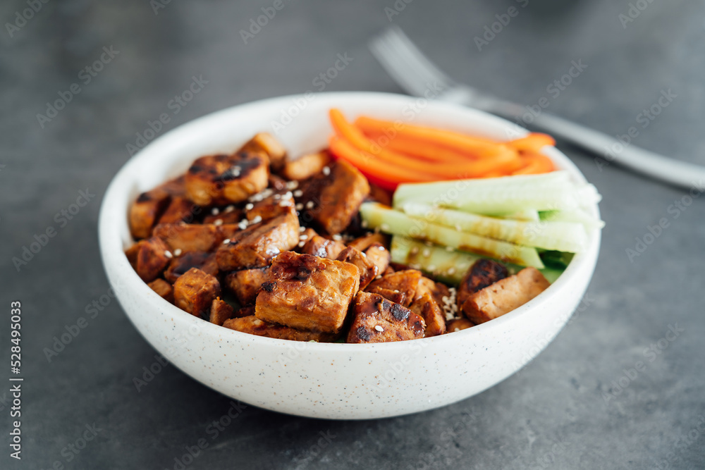 Bowl with roasted tempeh made of fermented soy beans, fresh vegetable sticks with sauce and sesame seeds on the table. Plant based protein. Healthy cooking and eating. Go vegan. selective focus Bowl with roasted tempeh made of fermented soy beans, fresh vegetable sticks with sauce and sesame seeds on the table. Plant based protein. Healthy cooking and eating. Go vegan. selective focus