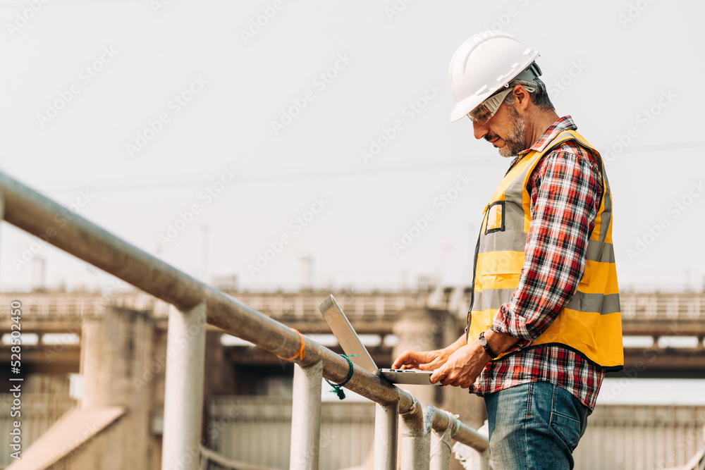 Portrait of hydropower engineer wearing safety jacket and hardhat with tablet working at outdoor field site that have water spillway  of hydro power dam electrical generator at the background.