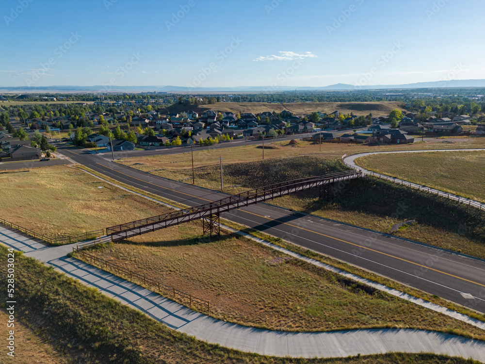 Footbridge for walking path and walkway crossing road with city scape and neighborhood in background with field grass during sunset in Wyoming from aerial drone