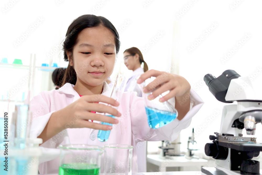Adorable Asian schoolgirl in lab coat doing science experiments, pouring blue reagent solution from beaker into test tube, happy smiling young scientist kid having fun during doing experiments, child Adorable Asian schoolgirl in lab coat doing science experiments, pouring blue reagent solution from beaker into test tube, happy smiling young scientist kid having fun during doing experiments, child