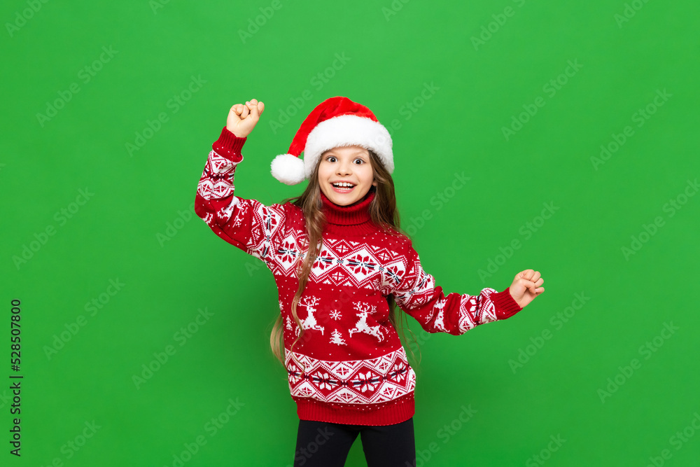 A little girl in a red sweater with reindeer and a Santa Claus hat waiting for the holiday on a green isolated background. The concept of Christmas. A little girl in a red sweater with reindeer and a Santa Claus hat waiting for the holiday on a green isolated background. The concept of Christmas.