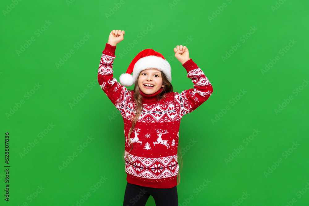 A little girl in a red sweater with reindeer and a Santa Claus hat waiting for the holiday on a green isolated background. The concept of Christmas. A little girl in a red sweater with reindeer and a Santa Claus hat waiting for the holiday on a green isolated background. The concept of Christmas.