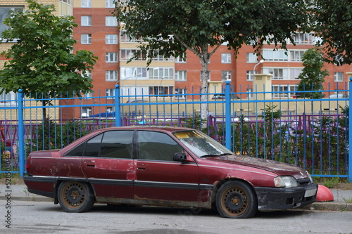 An old broken rusty abandoned dark red car stands near the blue fence, Soyuzny Prospekt, St. Petersburg, Russia, September 2022 © Станислав Вершинин