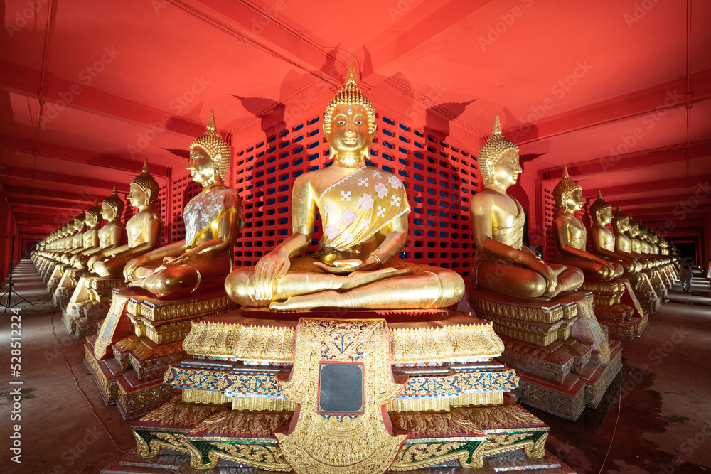 Several golden statues of Buddha statues in meditation posture are lined up in a corridor and spotlight on every statues in front of the red wall.