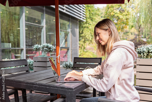 Young Caucasian business woman with blonde hair working on laptop in outdoor cafe. College student using technology , online education, freelance 
