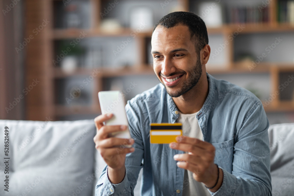 Online Shopping. Handsome Black Man Using Smartphone And Credit Card At Home