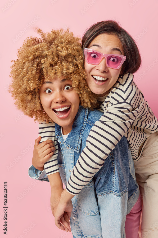 Positive happy women of different race have fun together look gladfully and surprised dressed in casual clothes isolated over pink background. Two female friends embrace foolish around indoors Positive happy women of different race have fun together look gladfully and surprised dressed in casual clothes isolated over pink background. Two female friends embrace foolish around indoors
