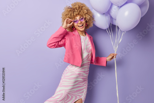 Indoor shot of stylish young woman wears long dress pink jacket and sunglasses holds bunch of inflated balloons enjoys party time isolated over purple background. People and holiday concept.
