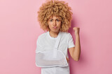 Angry irritated young woman with curly hair clenches fist feels annoyed wears sling in broken arm pouts lips isolated over pink background. Displeased female model has injury after accident.