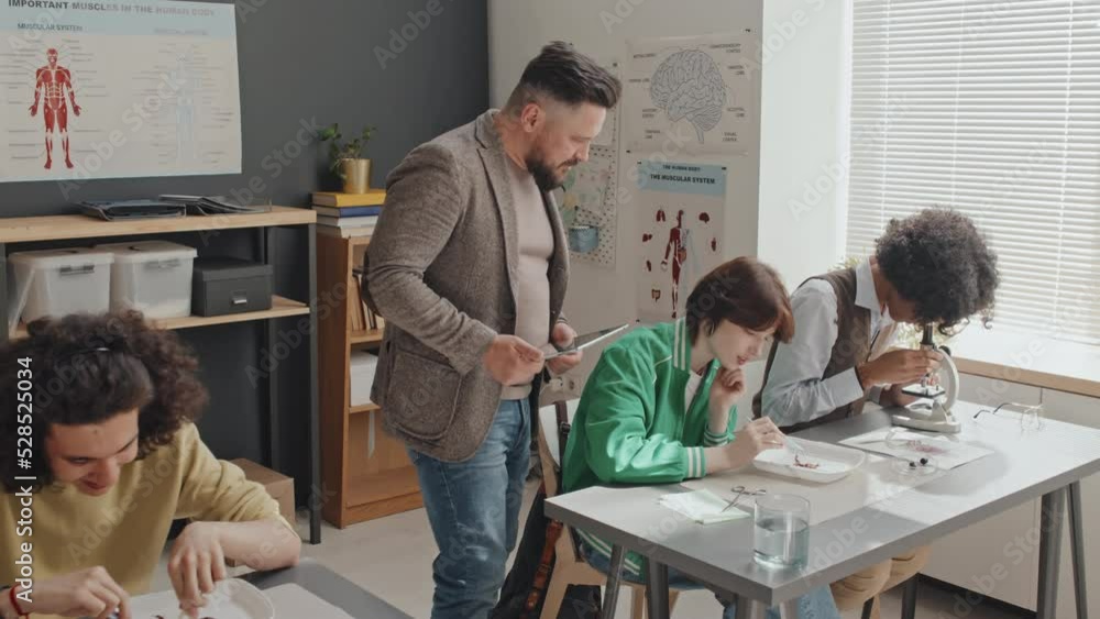 Medium long of bearded male teacher walking in classroom, watching diverse high school students sitting at desks, using microscopes and dissecting frogs Medium long of bearded male teacher walking in classroom, watching diverse high school students sitting at desks, using microscopes and dissecting frogs