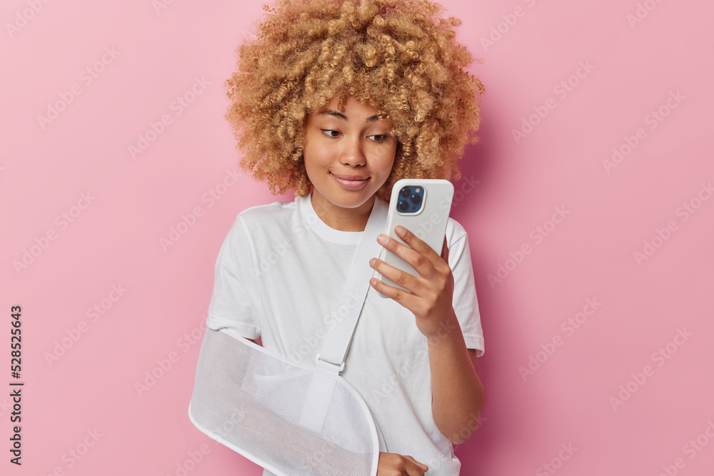 Positive good looking woman with curly bushy hair has injured arm in sling scrolls social networks focused at smartphone display with satisfied expression poses indoor against pink background Positive good looking woman with curly bushy hair has injured arm in sling scrolls social networks focused at smartphone display with satisfied expression poses indoor against pink background