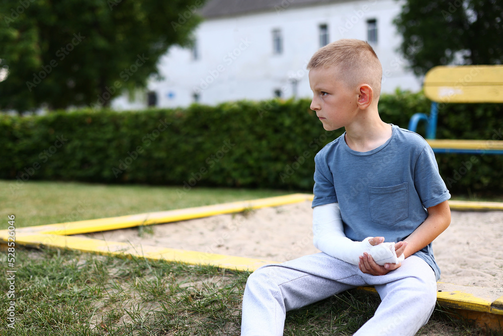 Fototapeta premium child caucasian with broken limb outdoors sits near the playground and looking on the plaster bandage on his arm. the worst summer vacation. concept of health accident, and medical